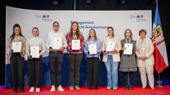 Acht Personen stehen nebeneinander auf einer Bühne mit rotem Teppich vor einem Hintergrund mit dem Logo und Schriftzug des Ministerpräsidenten von Schleswig-Holstein („Schleswig-Holstein – Der Ministerpräsident“). Alle tragen weiße Bänder mit Namensschildern um den Hals und halten Urkunden sowie kleine schwarze Etuis in den Händen. Sie blicken in die Kamera.  Die vierte Person von rechts ist Larissa Semelka, und die rechte Person ist Dr. Sabine Sütterlin-Waack. Rechts im Bild steht außerdem die Flagge von Schleswig-Holstein.