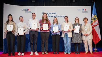 Acht Personen stehen nebeneinander auf einer Bühne mit rotem Teppich vor einem Hintergrund mit dem Logo und Schriftzug des Ministerpräsidenten von Schleswig-Holstein („Schleswig-Holstein – Der Ministerpräsident“). Alle tragen weiße Bänder mit Namensschildern um den Hals und halten Urkunden sowie kleine schwarze Etuis in den Händen. Sie blicken in die Kamera.  Die vierte Person von rechts ist Larissa Semelka, und die rechte Person ist Dr. Sabine Sütterlin-Waack. Rechts im Bild steht außerdem die Flagge von Schleswig-Holstein.