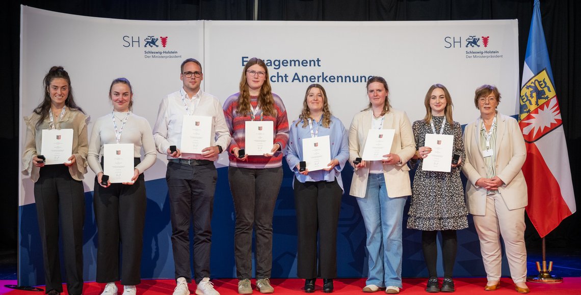 Acht Personen stehen nebeneinander auf einer Bühne mit rotem Teppich vor einem Hintergrund mit dem Logo und Schriftzug des Ministerpräsidenten von Schleswig-Holstein („Schleswig-Holstein – Der Ministerpräsident“). Alle tragen weiße Bänder mit Namensschildern um den Hals und halten Urkunden sowie kleine schwarze Etuis in den Händen. Sie blicken in die Kamera.  Die vierte Person von rechts ist Larissa Semelka, und die rechte Person ist Dr. Sabine Sütterlin-Waack. Rechts im Bild steht außerdem die Flagge von Schleswig-Holstein.